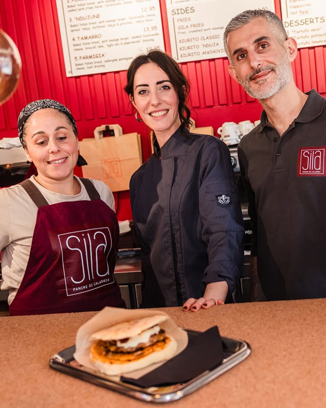 Raffaele, Carolina and Alba behind the counter of Sila, with the red interior and menu board